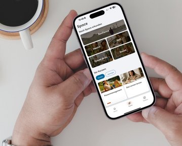 Ein Smartphone in der Hand zeigt eine App mit Kategorien wie Gesundheit, Selbst und Kurse. Im Hintergrund steht eine Tasse Kaffee.