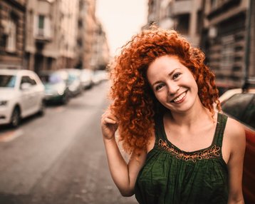 Eine Frau mit lockigem, rotem Haar lächelt und steht auf einer Stadtstraße, umgeben von parkenden Autos und Gebäuden. Sie trägt ein grünes Oberteil und hat eine entspannte Pose.