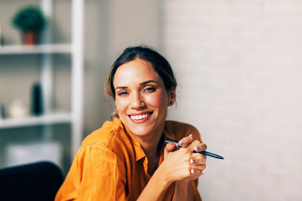 Eine lächelnde Frau mit einem Stift in der Hand sitzt an einem Schreibtisch. Im Hintergrund sind Regale mit verschiedenen Gegenständen und Pflanzen zu sehen. Sie trägt ein orangefarbenes Hemd.
