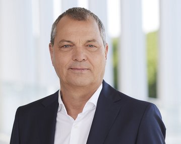 Mann in Anzug mit kurzem, grauen Haar, steht vor einem modernen Hintergrund mit großen Fenstern und grünem Ausblick.