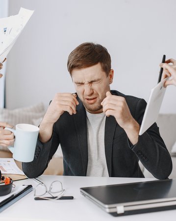 Ein Mann sitzt an einem Tisch, hält sich den Kopf und sieht gestresst aus, während Hände ihm Dokumente und eine Tasse reichen.
