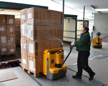 Ein Arbeiter transportiert mit einem elektrischen Hubwagen eine Palette mit Kartons aus einem Lieferwagen. Der Bereich ist eine Ladezone mit einem flachen Boden und weiteren Hubwagen im Hintergrund.