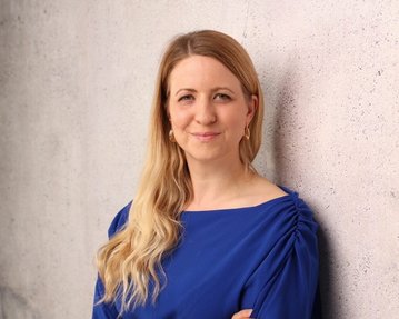 Eine Frau mit langen, blonden Haaren trägt ein blaues Oberteil und steht lächelnd an einer grauen Wand.