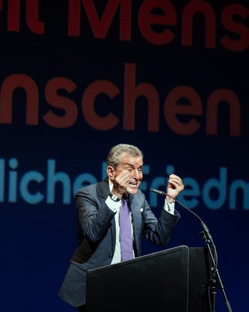 Ein Redner steht an einem Podium und gestikuliert während einer Veranstaltung. Im Hintergrund sind die Worte „Weil Menschen Menschen sind“ und „Mit Michael Friedman“ sichtbar.