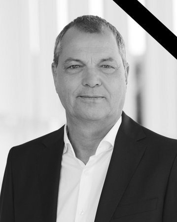 Porträt eines Mannes in einem schwarzen Anzug mit weißem Hemd, der direkt in die Kamera schaut. Ein Trauerrand ist im Bild vorhanden.