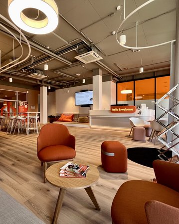 Moderne Lounge mit gemütlichen Sitzgelegenheiten, einem Tisch und einem Empfangsbereich. Ein Bildschirm zeigt "hello & welcome".