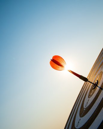 Eine Dartspitze steckt in der Mitte einer Zielscheibe mit konzentrischen Ringen, beleuchtet von Sonnenlicht im Hintergrund.