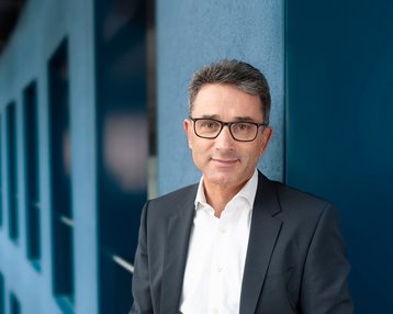 Ein Mann in einem Anzug steht an einer Wand in einem modernen Bürogebäude mit großen Fenstern und geschwungener Architektur.