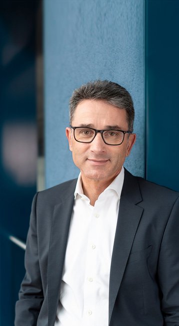 Ein Mann in einem Anzug steht an einer Wand in einem modernen Bürogebäude mit großen Fenstern und geschwungener Architektur.