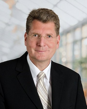 Ein Mann in einem schwarzen Anzug mit einer weißen Krawatte und Brille, der in einem hellen, modernen Raum steht. Der Hintergrund zeigt unscharfe Pflanzen und Fenster.