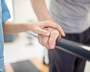Eine Hand hält eine Stange, während eine andere Hand Unterstützung bietet. Die Szene zeigt eine therapeutische Umgebung.