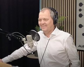 Ein Mann mit Kopfhörern steht vor einem Mikrofon in einem Studio, lächelnd, mit einem weißen Hemd und einer Tasse auf dem Tisch.