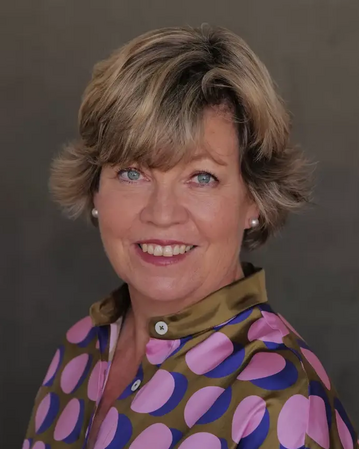 Eine Frau mit blonden, lockigen Haaren trägt ein buntes Hemd mit großen Punkten und lächelt freundlich in die Kamera.