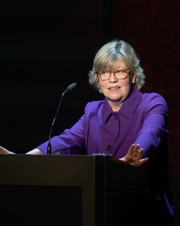 Eine Person mit blonden Haaren und Brille steht an einem Rednerpult und spricht, gekleidet in einem lila Oberteil.