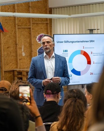 Ein Mann in einem Anzug spricht vor einem Publikum, während eine Präsentation mit Diagrammen und Statistiken im Hintergrund angezeigt wird. Die Zuschauer halten Smartphones und hören aufmerksam zu.