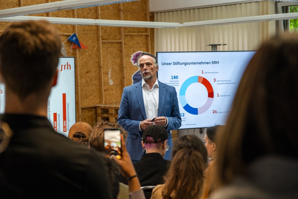 Ein Mann in einem Anzug spricht vor einem Publikum, während eine Präsentation mit Diagrammen und Statistiken im Hintergrund angezeigt wird. Die Zuschauer halten Smartphones und hören aufmerksam zu.