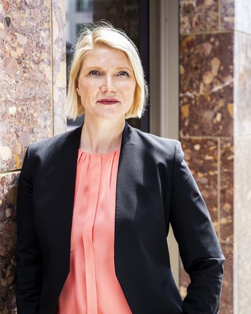 Eine Frau mit blonden, kurzen Haaren trägt ein schwarzes Blazer und ein rosa Oberteil. Sie steht an einer Wand aus Marmor und schaut direkt in die Kamera.