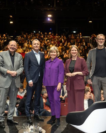Fünf Personen stehen auf einer Bühne vor einem Publikum in einem Theater. Sie lächeln und tragen formelle Kleidung.