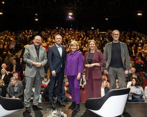 Fünf Personen stehen auf einer Bühne vor einem Publikum in einem Theater. Sie lächeln und tragen formelle Kleidung.