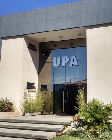 Eingang eines modernen Gebäudes mit der Aufschrift "UPA". Vor dem Gebäude befinden sich Pflanzen und Flaggen. Der Eingang ist verglast und gut beleuchtet.
