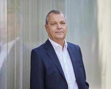 Prof. Dr. Christof Hettich in einem dunklen Anzug steht an einer Wand aus transparenten Materialien und schaut direkt in die Kamera.