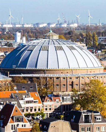 Eine große, runde Halle mit einem gewölbten Dach steht im Vordergrund, umgeben von städtischen Gebäuden und Windkraftanlagen im Hintergrund.