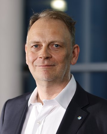 Ein Mann mit kurzen, grauen Haaren und einem leichten Bart trägt einen schwarzen Anzug und ein weißes Hemd.