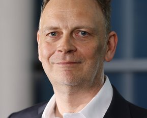 Ein Mann mit kurzen, grauen Haaren und einem leichten Bart trägt einen schwarzen Anzug und ein weißes Hemd.