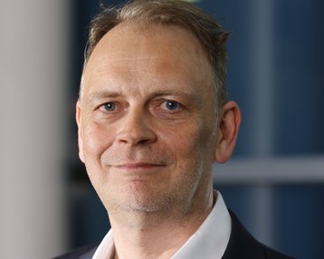 Ein Mann mit kurzen, grauen Haaren und einem leichten Bart trägt einen schwarzen Anzug und ein weißes Hemd.