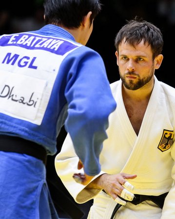 Zwei Judokas stehen sich gegenüber, bereit für den Wettkampf. Der Kämpfer links trägt einen blauen Judogi mit der Aufschrift "E. Batzava" und "MGL", während der Kämpfer rechts einen weißen Judogi trägt.