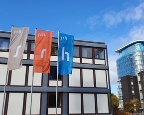 Drei Fahnen mit den Buchstaben "s", "r" und "h" stehen vor einem modernen Gebäude mit Glasfassade. Im Hintergrund ist ein weiterer moderner Bau sichtbar. Der Himmel ist teilweise bewölkt.