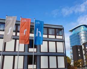 Drei Fahnen mit den Buchstaben "s", "r" und "h" stehen vor einem modernen Gebäude mit Glasfassade. Im Hintergrund ist ein weiterer moderner Bau sichtbar. Der Himmel ist teilweise bewölkt.