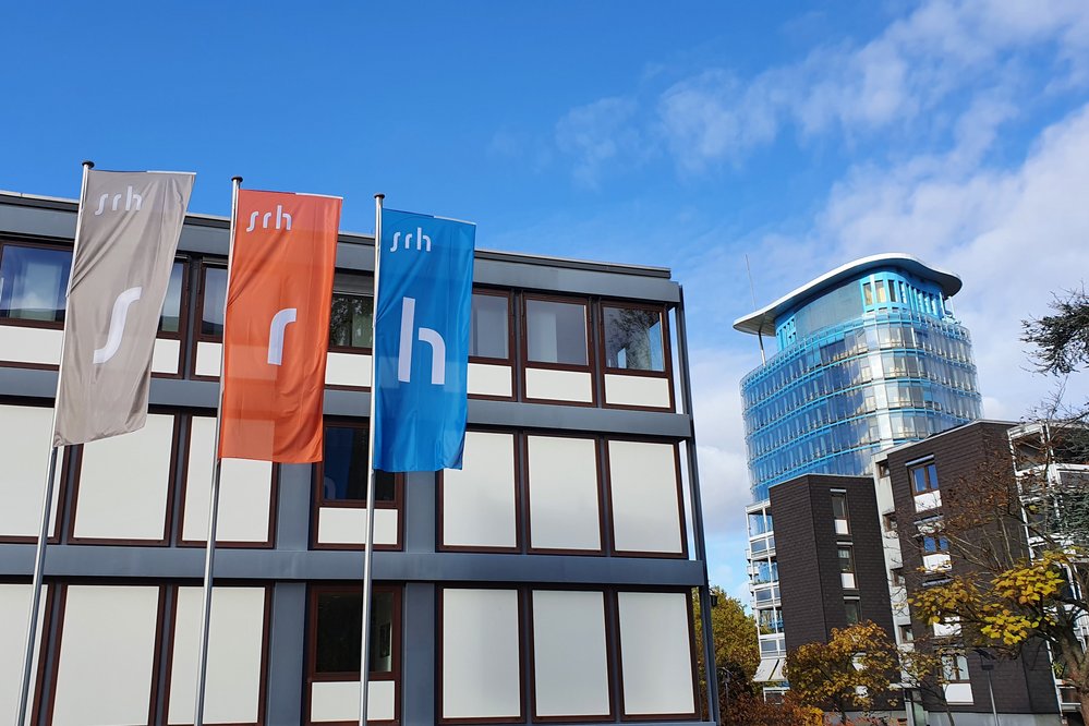Drei Fahnen mit den Buchstaben "s", "r" und "h" stehen vor einem modernen Gebäude mit Glasfassade. Im Hintergrund ist ein weiterer moderner Bau sichtbar. Der Himmel ist teilweise bewölkt.