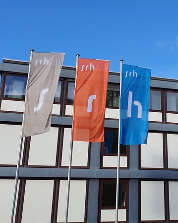 Drei Fahnen mit den Buchstaben "s", "r" und "h" stehen vor einem modernen Gebäude mit Glasfassade. Im Hintergrund ist ein weiterer moderner Bau sichtbar. Der Himmel ist teilweise bewölkt.
