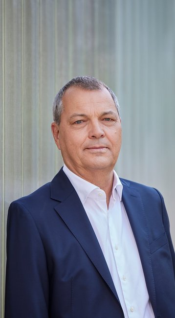 Ein Mann in einem dunklen Anzug steht vor einer transparenten Wand. Er blickt direkt in die Kamera.