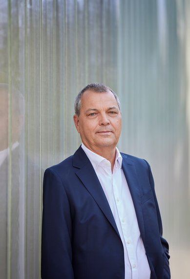 Ein Mann in einem dunklen Anzug steht vor einer transparenten Wand. Er blickt direkt in die Kamera.