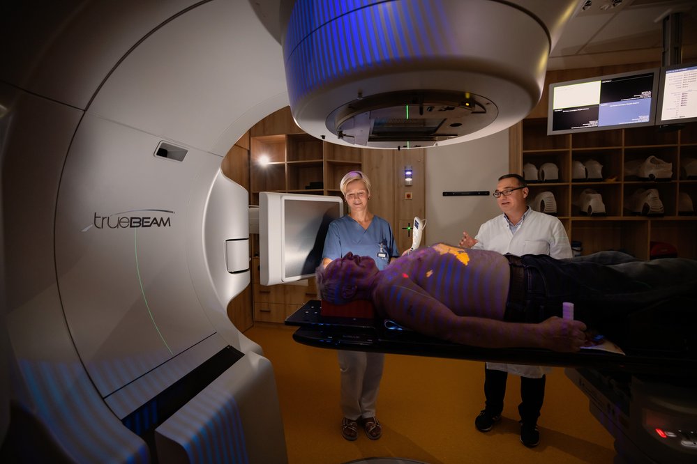 Ein Patient liegt auf einer Behandlungsliege unter einem TrueBeam-Gerät, während zwei medizinische Fachkräfte ihn vorbereiten. Das Gerät ist für die Strahlentherapie konzipiert.