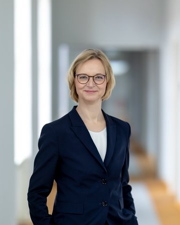 Eine Frau mit blonden Haaren und Brille trägt einen dunkelblauen Blazer. Sie steht in einem hellen Flur mit mehreren Türen im Hintergrund und lächelt leicht.