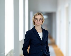 Eine Frau mit blonden Haaren und Brille trägt einen dunkelblauen Blazer. Sie steht in einem hellen Flur mit mehreren Türen im Hintergrund und lächelt leicht.