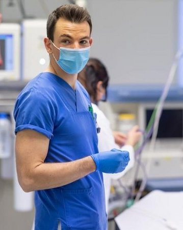 Ein Arzt in blauer Kleidung und Maske steht an einem Bett in einem Krankenhaus, während zwei weitere medizinische Fachkräfte im Hintergrund arbeiten. Alle tragen Schutzmasken und sind mit medizinischen Geräten umgeben.