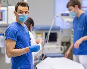 Ein Arzt in blauer Kleidung und Maske steht an einem Bett in einem Krankenhaus, während zwei weitere medizinische Fachkräfte im Hintergrund arbeiten. Alle tragen Schutzmasken und sind mit medizinischen Geräten umgeben.