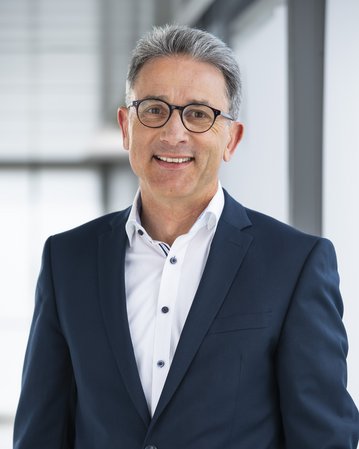 Mann in einem dunkelblauen Anzug mit Brille, lächelnd vor einem modernen Hintergrund.