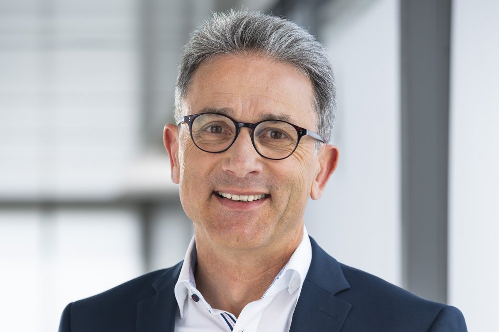 Mann in einem dunkelblauen Anzug mit Brille, lächelnd vor einem modernen Hintergrund.