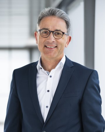 Foto: Ein Mann mit Brille und grauen Haaren trägt einen dunkelblauen Anzug und ein weißes Hemd. Er lächelt freundlich vor einem modernen Hintergrund.