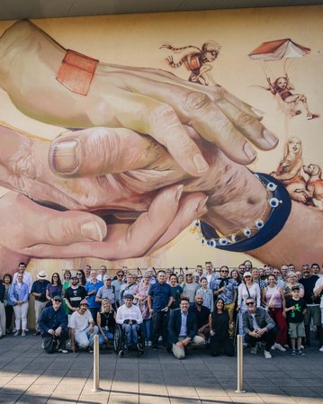 Eine große Gruppe von Menschen steht vor einem Wandgemälde, das zwei Hände zeigt, die sich berühren, umgeben von verschiedenen Figuren.