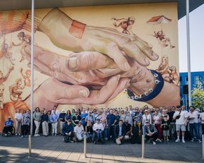 Eine große Gruppe von Menschen steht vor einem Wandgemälde, das zwei Hände zeigt, die sich berühren, umgeben von verschiedenen Figuren.