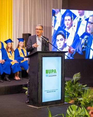 Patrick Mombaur steht an einem Podium während der UPA-Abschlussfeier, im Hintergrund sind Absolventen in blauen Roben und Hüten zu sehen.