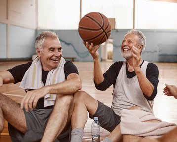 Vier ältere Männer sitzen auf dem Boden eines Basketballfeldes, während einer von ihnen einen Basketball in der Hand hält und lächelt. Die anderen schauen ihn an und scheinen am Gespräch beteiligt zu sein.