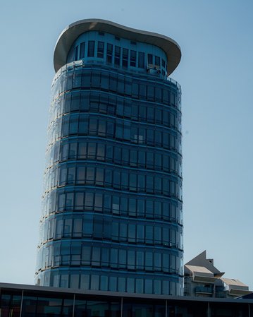 Moderne Glasfassade eines hohen Gebäudes mit abgerundetem Dach, umgeben von anderen Gebäuden und klarem Himmel.