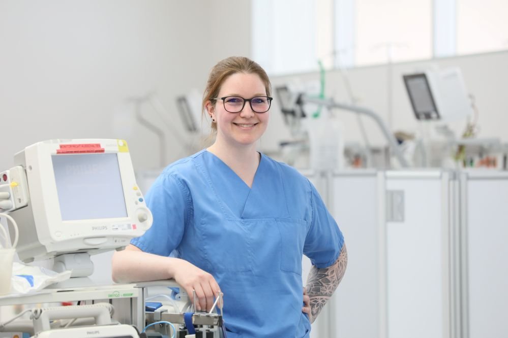 Eine Krankenschwester in blauer Arbeitskleidung steht lächelnd neben medizinischen Geräten in einem modernen Krankenhausumfeld.